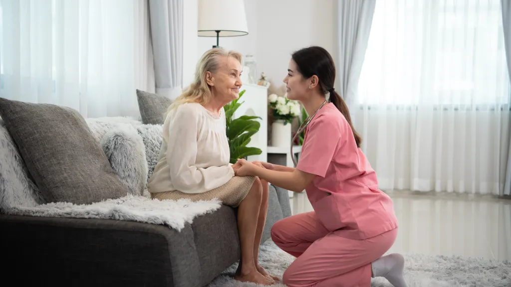 Home care aide kneeling to talk with an older woman in her living room